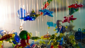 Hanging glass sculptures depicting various shapes and colors are suspended on thin strings. The glass pieces are intricately designed with vibrant colors like blue, red, and green, creating a playful cacophony as they catch the light. The arrangement provides a sense of depth and whimsy.