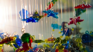 Hanging glass sculptures depicting various shapes and colors are suspended on thin strings. The glass pieces are intricately designed with vibrant colors like blue, red, and green, creating a playful cacophony as they catch the light. The arrangement provides a sense of depth and whimsy.