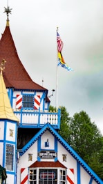 A building with distinct Bavarian-style architecture, featuring white walls with blue wooden trim and red accents. The roof is steep and brown, adorned with ornamental features. Two flags on poles are present; one is the United States flag, while the other resembles a Bavarian flag.