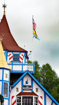 A building with distinct Bavarian-style architecture, featuring white walls with blue wooden trim and red accents. The roof is steep and brown, adorned with ornamental features. Two flags on poles are present; one is the United States flag, while the other resembles a Bavarian flag.