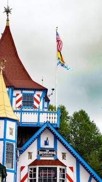 A building with distinct Bavarian-style architecture, featuring white walls with blue wooden trim and red accents. The roof is steep and brown, adorned with ornamental features. Two flags on poles are present; one is the United States flag, while the other resembles a Bavarian flag.