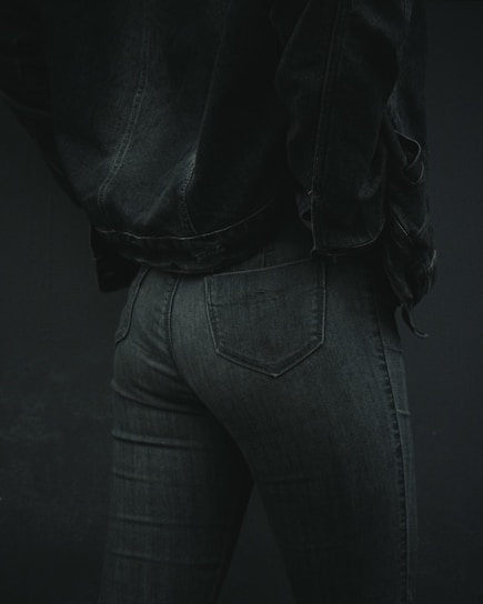 A person is wearing dark denim jeans and a matching denim jacket. The focus is on the lower back and hip area, showcasing the texture and fit of the clothing. The lighting is low, emphasizing shadows and details in the fabric.