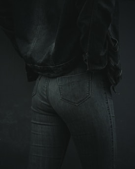 A person is wearing dark denim jeans and a matching denim jacket. The focus is on the lower back and hip area, showcasing the texture and fit of the clothing. The lighting is low, emphasizing shadows and details in the fabric.