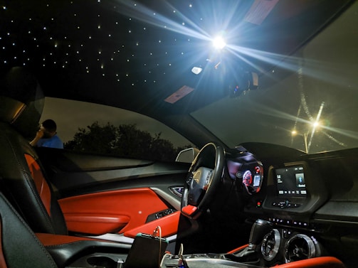 A luxurious car interior featuring red and black leather seats, illuminated dashboard, and a ceiling designed to resemble a starry night. The ambient lighting creates a futuristic and sophisticated atmosphere. Outside the car window, a person stands talking on the phone next to some trees.