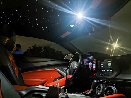 A luxurious car interior featuring red and black leather seats, illuminated dashboard, and a ceiling designed to resemble a starry night. The ambient lighting creates a futuristic and sophisticated atmosphere. Outside the car window, a person stands talking on the phone next to some trees.