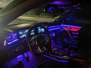 The interior of a modern car illuminated with ambient lighting. The dashboard features a digital display with multiple gauges and controls. The steering wheel is visible, along with various control buttons on it. The ambient lighting consists of blue and purple hues that accentuate the car's futuristic design, visible on the dashboard and the passenger side.