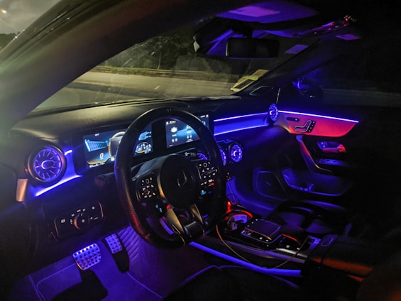 The interior of a modern car illuminated with ambient lighting. The dashboard features a digital display with multiple gauges and controls. The steering wheel is visible, along with various control buttons on it. The ambient lighting consists of blue and purple hues that accentuate the car's futuristic design, visible on the dashboard and the passenger side.