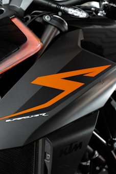 A close-up view of a motorcycle part featuring sleek black and orange design elements. The bodywork showcases the word 'SUPERDUKE' in stylized white text. The logo 'KTM' is also visible, indicating the motorcycle's brand. The image focuses on the angular lines and the modern, aggressive styling of the vehicle.