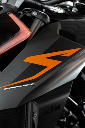 A close-up view of a motorcycle part featuring sleek black and orange design elements. The bodywork showcases the word 'SUPERDUKE' in stylized white text. The logo 'KTM' is also visible, indicating the motorcycle's brand. The image focuses on the angular lines and the modern, aggressive styling of the vehicle.
