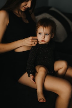 A young child with light skin and light brown hair, dressed in a dark, patterned outfit, is sitting on an adult's lap. The adult, with long brown hair, is gently holding the child. The setting is indoors, with dim lighting, giving a cozy and intimate feel.