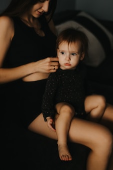 A young child with light skin and light brown hair, dressed in a dark, patterned outfit, is sitting on an adult's lap. The adult, with long brown hair, is gently holding the child. The setting is indoors, with dim lighting, giving a cozy and intimate feel.