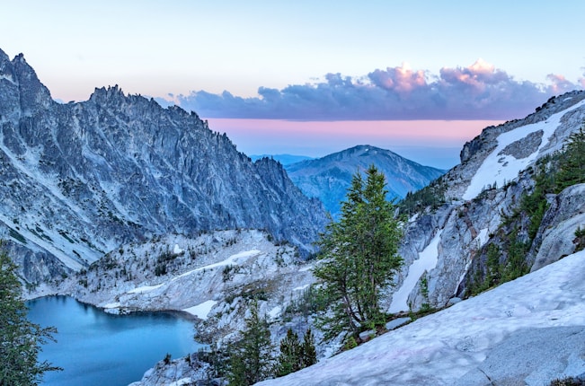 An alpine landscape with rugged, snow-dusted mountains surrounding a serene turquoise lake. Pines and rocky outcroppings populate the foreground, while the sky is painted in soft hues of pink and blue, with fluffy clouds scattered across.