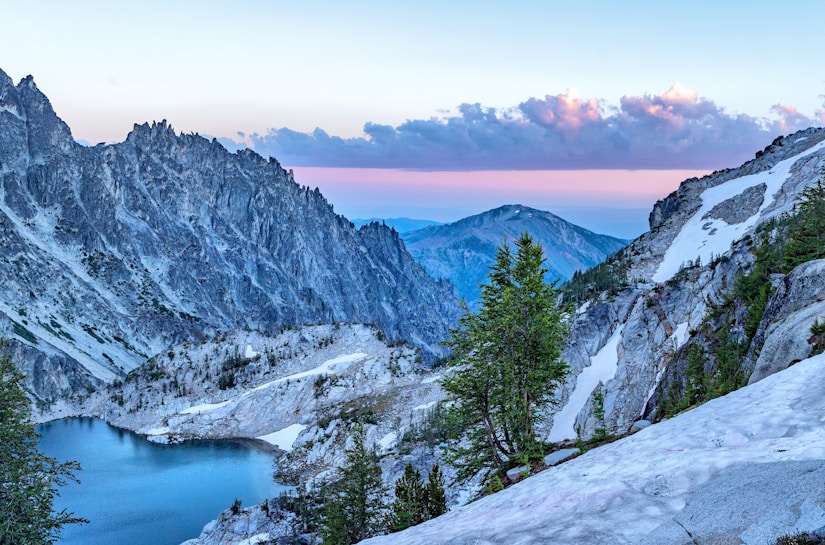 An alpine landscape with rugged, snow-dusted mountains surrounding a serene turquoise lake. Pines and rocky outcroppings populate the foreground, while the sky is painted in soft hues of pink and blue, with fluffy clouds scattered across.