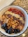 A bowl filled with a blend of ingredients including fresh blueberries, crunchy granola, cacao nibs, hemp seeds, and goji berries, all arranged on a layer of purple acai or smoothie blend. The mixture is presented in a white bowl with a black rim, set on a marbled surface.