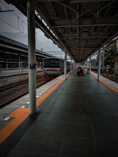 A train platform with an overhead roof structure, featuring a stationary train with people sitting on benches. The scene is captured in low light, giving an overcast and moody atmosphere. The platform is lined with orange tactile paving and flanked by train tracks on one side.