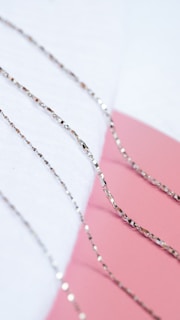 Several delicate, reflective silver chains are neatly arranged on a soft white surface, with a contrasting pink background creating a visually appealing composition.