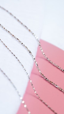 Several delicate, reflective silver chains are neatly arranged on a soft white surface, with a contrasting pink background creating a visually appealing composition.