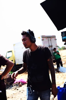 A person wearing headphones stands outdoors in a bright, sunny location. They have a casual posture and appear to be listening intently or focused. In the background, there are trucks and other people, suggesting a work or event setting.