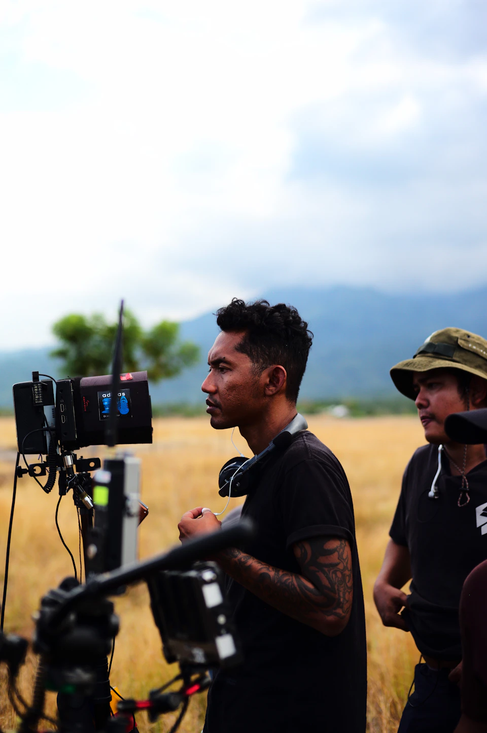 The image shows a man standing on a film set, holding a camera and directing it towards another person who is also present on the set. The man with the camera appears to be focused on capturing the scene, while his companion stands nearby. In the background, there are mountains visible in the distance, adding depth to the scene.