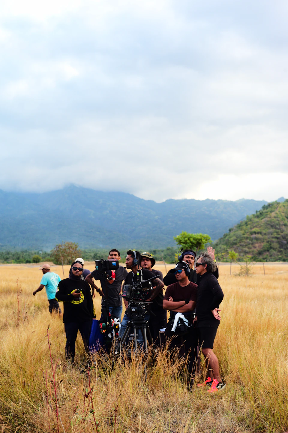 Group of people stand on grassy field, surrounded by mountains under overcast sky outdoors