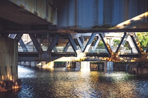 A detailed view of the underside of a bridge with water flowing beneath. Metal beams and girders form a complex, industrial structure. The sunlight creates shadows and highlights on the metallic surfaces, adding depth and contrast. Greenery is visible in the background, suggesting the presence of nature nearby.