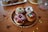 A selection of chocolate-glazed donuts sprinkled with colorful decorations on a wooden plate. Additional donuts are placed in the background on a white platter, with assorted sprinkles scattered on a wooden table. A glass bowl with more sprinkles is partially visible, creating a joyful and festive atmosphere.