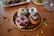 A selection of chocolate-glazed donuts sprinkled with colorful decorations on a wooden plate. Additional donuts are placed in the background on a white platter, with assorted sprinkles scattered on a wooden table. A glass bowl with more sprinkles is partially visible, creating a joyful and festive atmosphere.