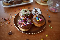 A selection of chocolate-glazed donuts sprinkled with colorful decorations on a wooden plate. Additional donuts are placed in the background on a white platter, with assorted sprinkles scattered on a wooden table. A glass bowl with more sprinkles is partially visible, creating a joyful and festive atmosphere.