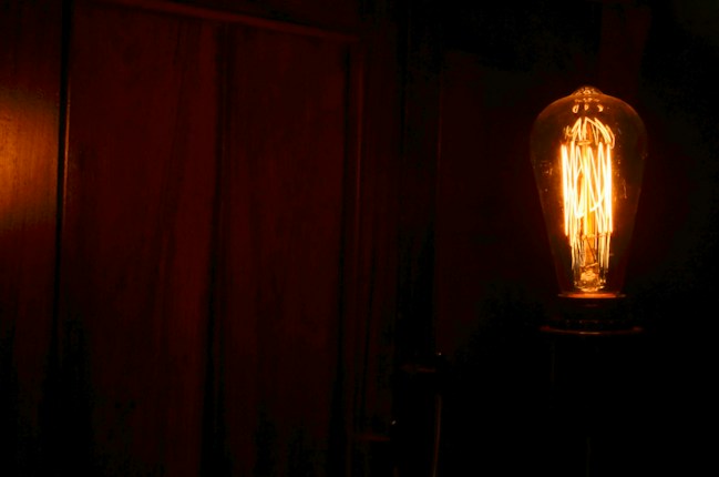 A dimly lit room features a vintage-style Edison bulb illuminated against a dark wooden backdrop. The filament inside the bulb glows warmly, casting a soft amber light.