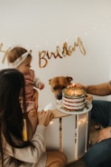 A family celebration scene features a baby in a pink dress held by a woman, a cake on a table with a single candle, and a dog curiously observing the cake. A gold 'Happy Birthday' banner is in the background.