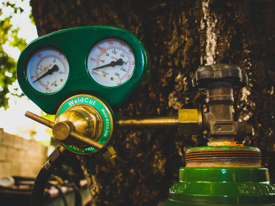 The image shows a green Welder's tool with two gauges attached to it, one on each side. The gauge on the left is larger and has a white face with black markings, while the gauge on the right is smaller and also features a white face with black markings.

The tool is resting against a tree trunk, which serves as a backdrop for this industrial scene.
