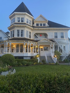 A large and ornate Victorian-style house adorned with string lights, featuring a prominent turret and multiple gabled roofs. The front porch is supported by white pillars, and there are well-maintained gardens with lush greenery in the foreground. The building has a sign indicating it is a restaurant.