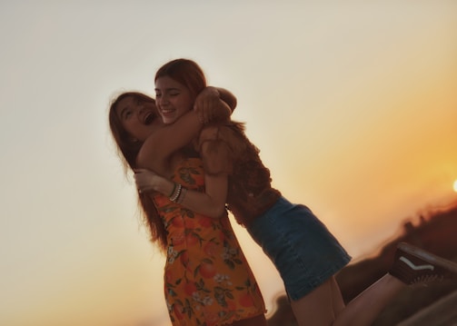 Two young women are joyfully embracing each other during a sunset. They are smiling and appear to be in a lively mood. One is wearing a floral dress, while the other is in a denim skirt and blouse. The scene conveys a sense of warmth and happiness.