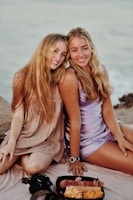Two individuals are sitting closely together on a beach, dressed in casual summer attire. They both have long hair and are smiling happily. A tray with snacks, including cheese and crackers, is placed in front of them on a blanket. The setting appears relaxed and inviting, with a calm ocean in the background.