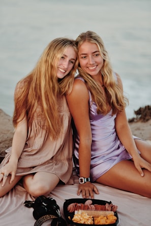 Two individuals are sitting closely together on a beach, dressed in casual summer attire. They both have long hair and are smiling happily. A tray with snacks, including cheese and crackers, is placed in front of them on a blanket. The setting appears relaxed and inviting, with a calm ocean in the background.