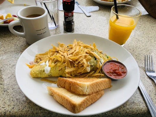 A breakfast table setting with a plate containing an omelette topped with shredded cheese and sour cream, accompanied by two slices of toast and a small dish of salsa. There's a glass of orange juice with a straw and a cup of coffee. A black pepper grinder and a glass of water are visible in the background.