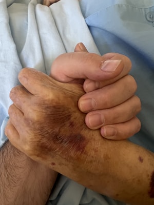 A close-up view of two hands clasped together, one appearing older with visible age spots and the other younger. They rest on a fabric that seems to be a light blue or white sheet or garment.