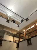 A multi-level interior with an open ceiling, showcasing architectural lines and a combination of wooden and metal railings. Industrial elements such as exposed pipes and lighting are visible. The word 'ANJUNG' is illuminated on a sign, and decorative wall art is present.