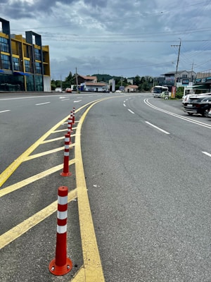 A curving road with red and white traffic cones lined along the yellow lane divider, leading to a commercial area with buildings on the left side. Several vehicles are visible, including parked cars and a truck moving along the street. Overhead, power lines stretch across the scene under a cloudy sky.