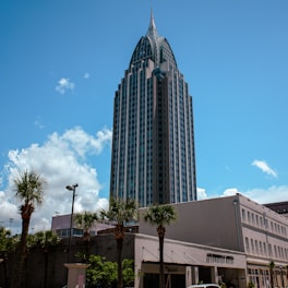 A tall, modern skyscraper with a pointed roof piercing the clear blue sky. In the foreground, several palm trees line the street and a smaller building stands nearby with a sign reading 'Arthur R. Outlaw Convention Center'. Fluffy white clouds dot the otherwise clear sky.