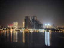 A nighttime cityscape featuring illuminated high-rise buildings reflecting off a calm body of water. The skyline is composed of modern architecture with bright lights creating a contrast against the dark night sky. The reflections on the water add a sense of tranquility to the scene.