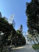 A modern cityscape featuring a geometrically designed skyscraper contrasted against lush greenery with a clear blue sky. A few people are walking along a paved path, near a white building with multiple windows. The scene combines urban architecture with a touch of nature.