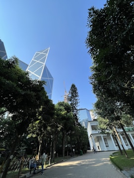 A modern cityscape featuring a geometrically designed skyscraper contrasted against lush greenery with a clear blue sky. A few people are walking along a paved path, near a white building with multiple windows. The scene combines urban architecture with a touch of nature.