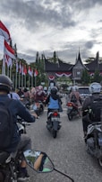 A group of people on motorbikes is gathered in front of a traditional building with a unique roof architecture, possibly in Indonesia. The scene is filled with national flags lining the street, creating a sense of patriotism and commemoration.