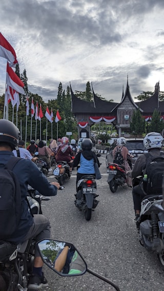 A group of people on motorbikes is gathered in front of a traditional building with a unique roof architecture, possibly in Indonesia. The scene is filled with national flags lining the street, creating a sense of patriotism and commemoration.