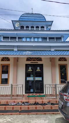 A building with a prominent blue-tiled dome and a flat roof below it. There are several windows with brown frames and a central doorway with the words 'DARUL AMAL' written on it. A variety of shoes are neatly placed on the stairs leading to the entrance, and a dark-colored vehicle is partially visible on the right side of the image.