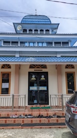 A building with a prominent blue-tiled dome and a flat roof below it. There are several windows with brown frames and a central doorway with the words 'DARUL AMAL' written on it. A variety of shoes are neatly placed on the stairs leading to the entrance, and a dark-colored vehicle is partially visible on the right side of the image.