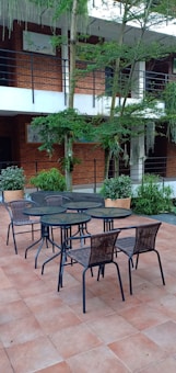A patio area with a tile floor featuring a set of black metal tables and wicker chairs. The background includes brick walls, lush greenery, and a tree growing through the middle. There are potted plants along the perimeter and railing on the upper floor of the building.