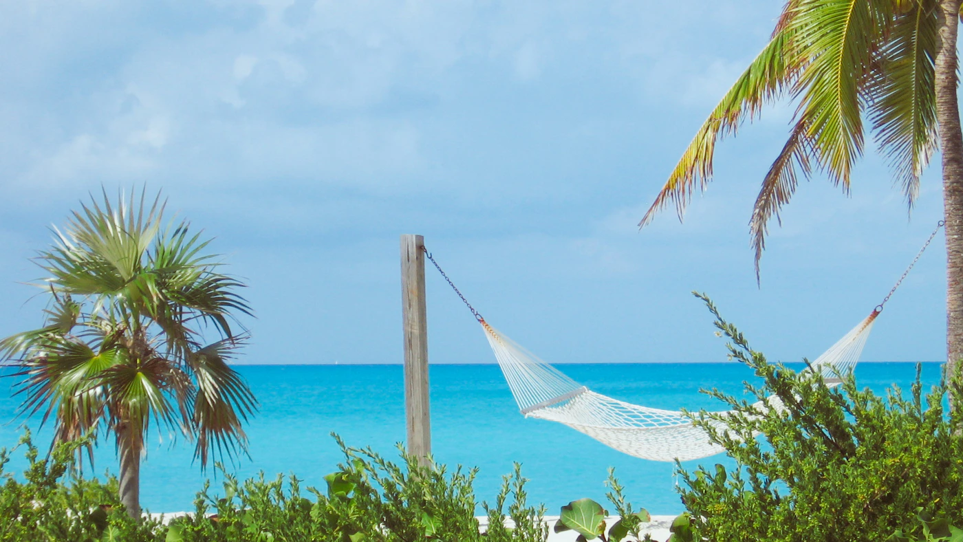 A hammock hanging between two palm trees on a white sand beach in the Bahamas with the turquoise Caribbean water visible beyond and a clear blue sky above