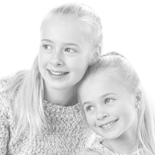Two young children with light hair are portrayed in a black and white image. They wear textured sweaters and have their hair tied back. The older child looks outward with a gentle smile, while the younger child leans into the older one, smiling warmly.